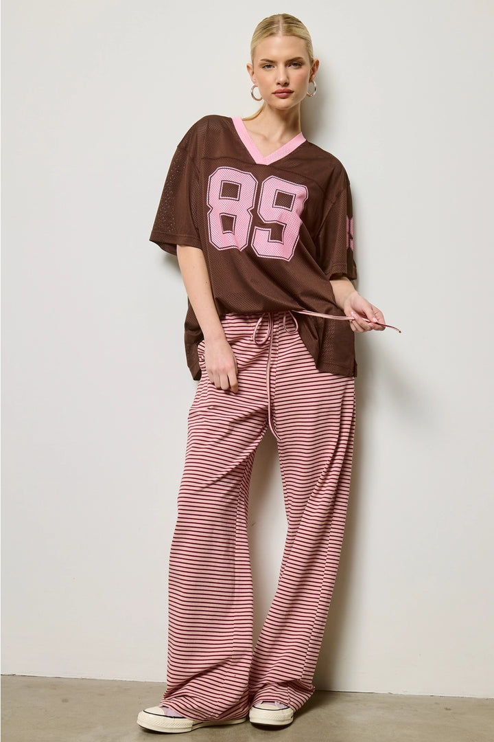 Woman wearing a brown oversized jersey with pink numbers and red striped pants against a white background