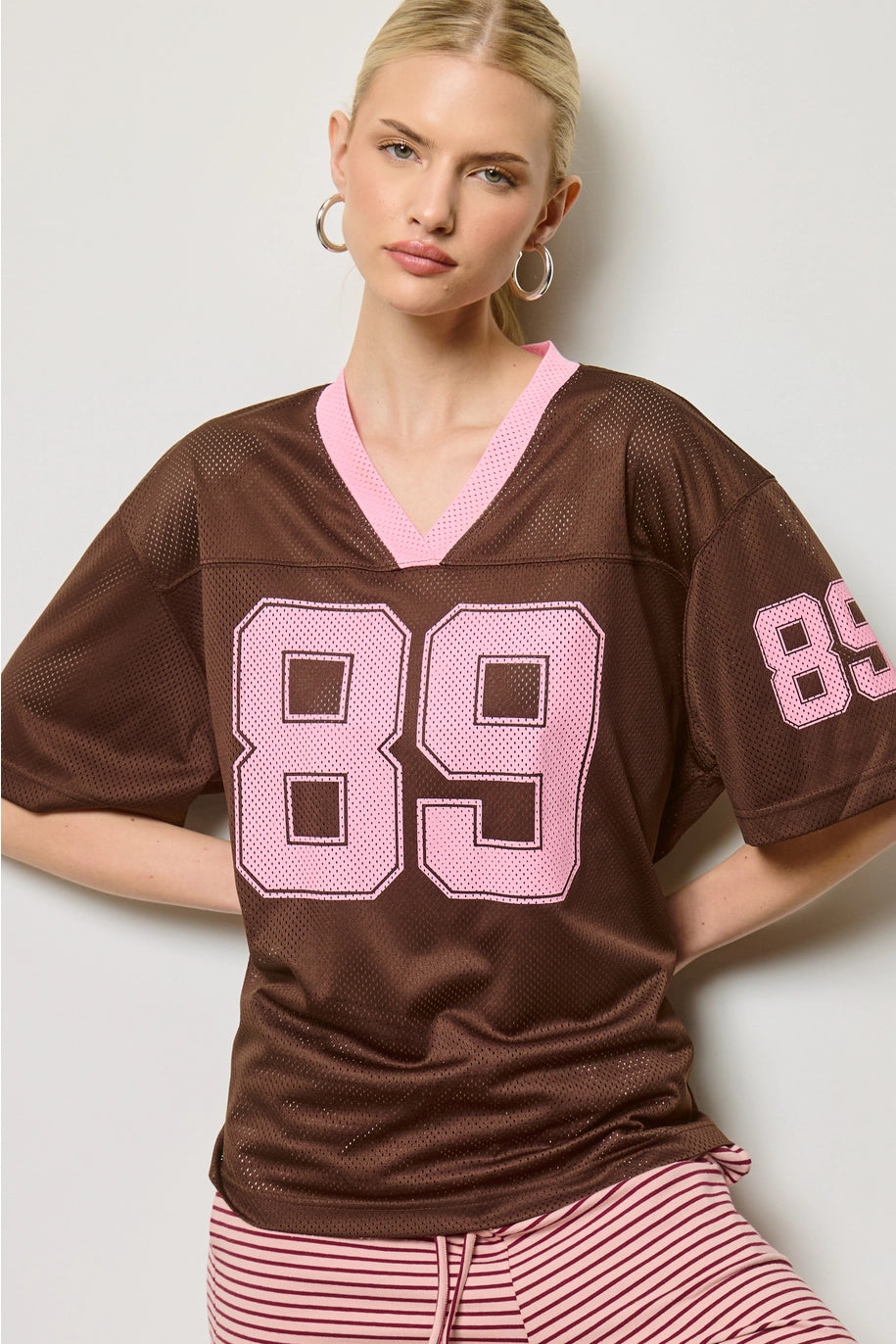 Woman wearing a brown jersey with pink numbers on a plain background