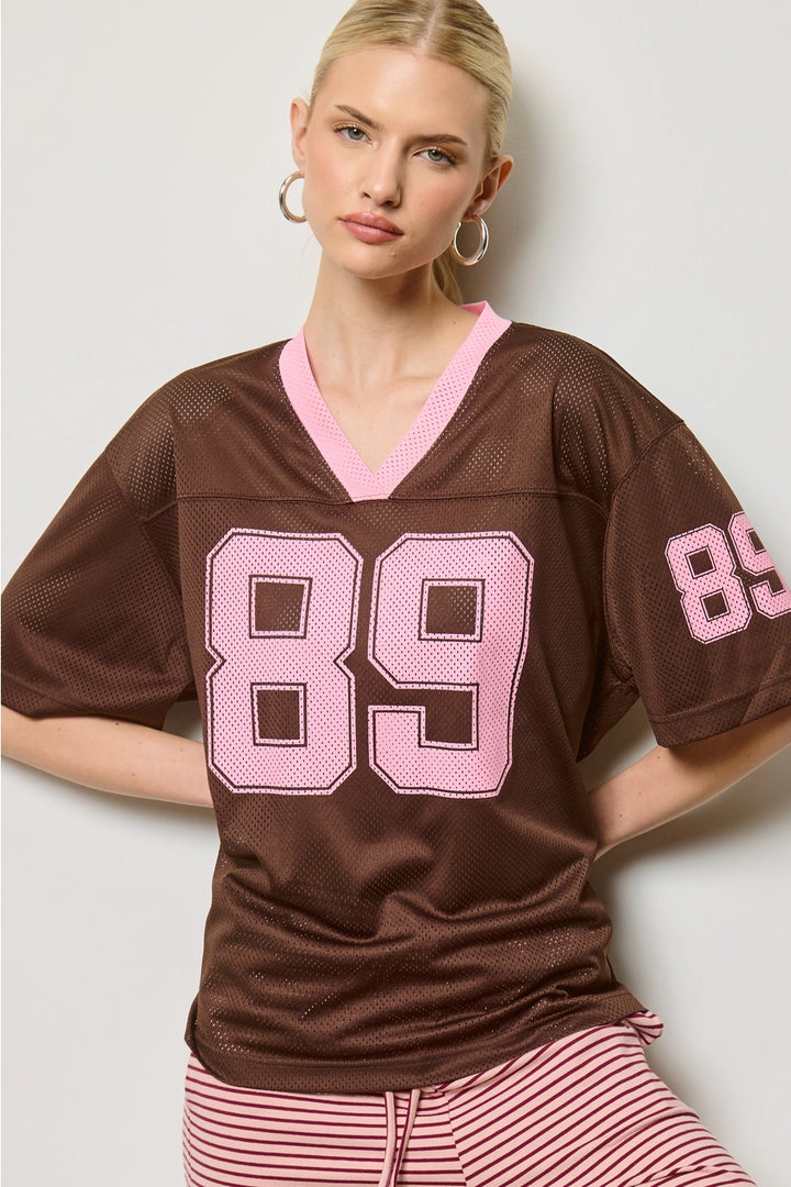 Woman wearing a brown jersey with pink numbers on a plain background