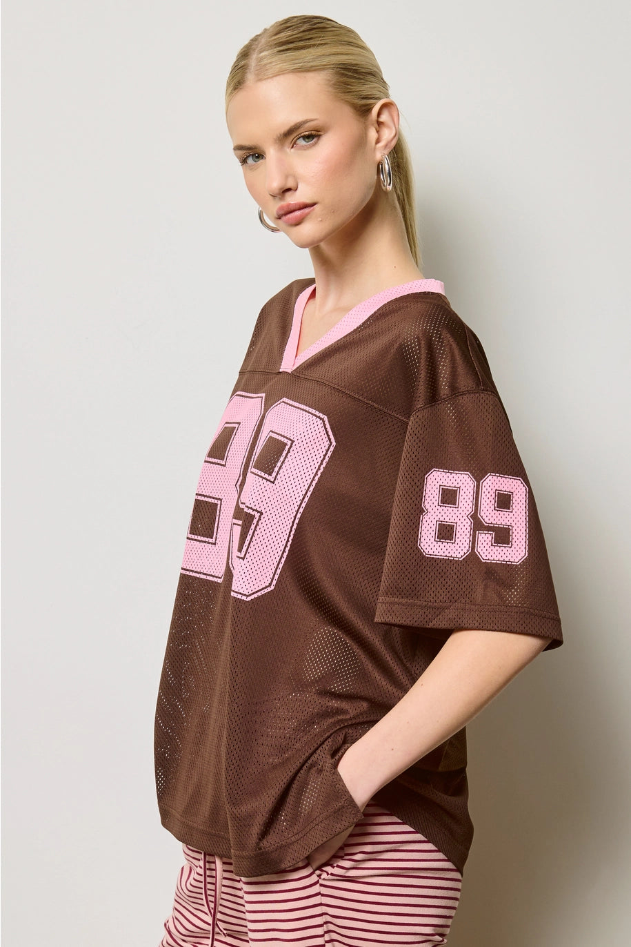 Woman wearing a brown jersey with pink numbers against a plain background