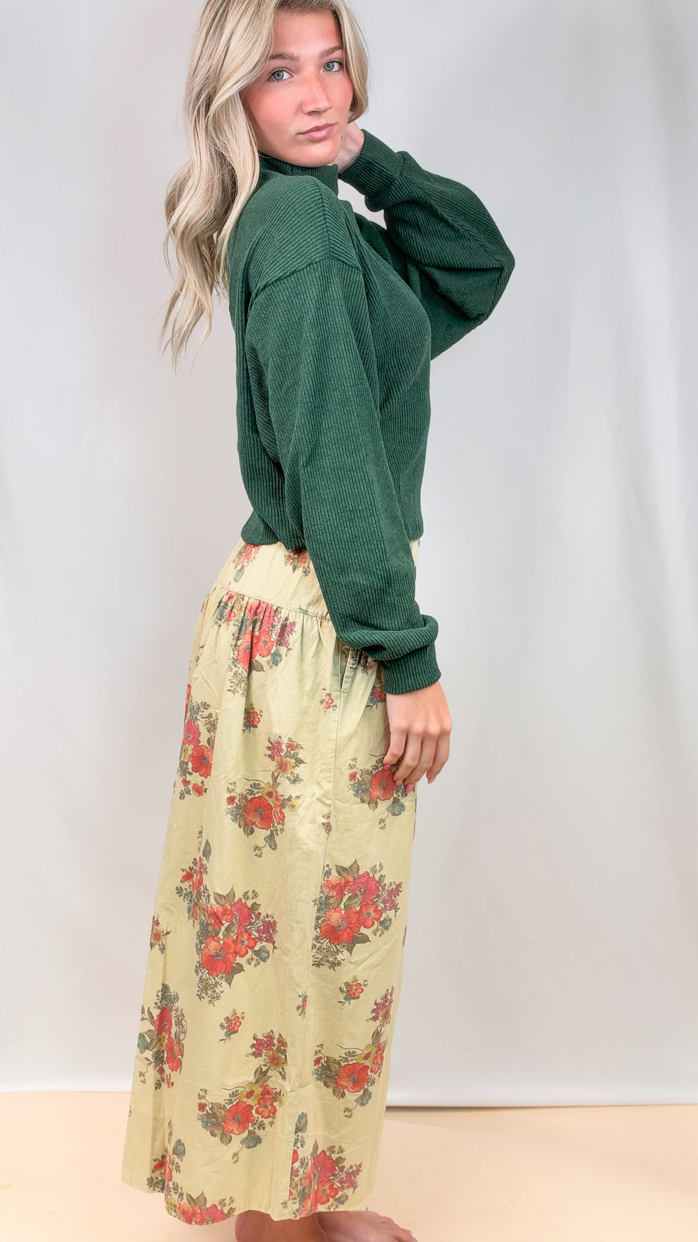 Woman shown from side view wearing green sweater and floral pants on plain background
