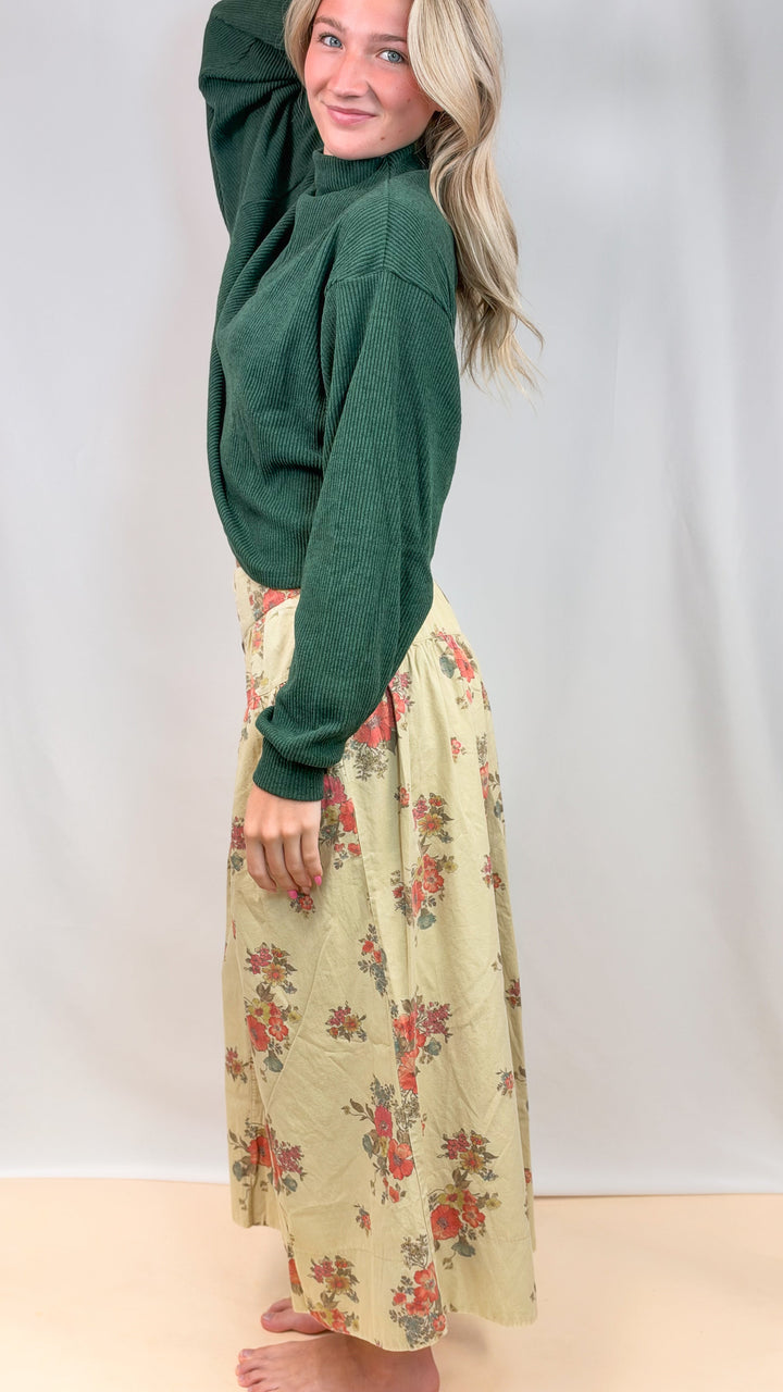 Woman shown from side view wearing green sweater with floral pants on plain background