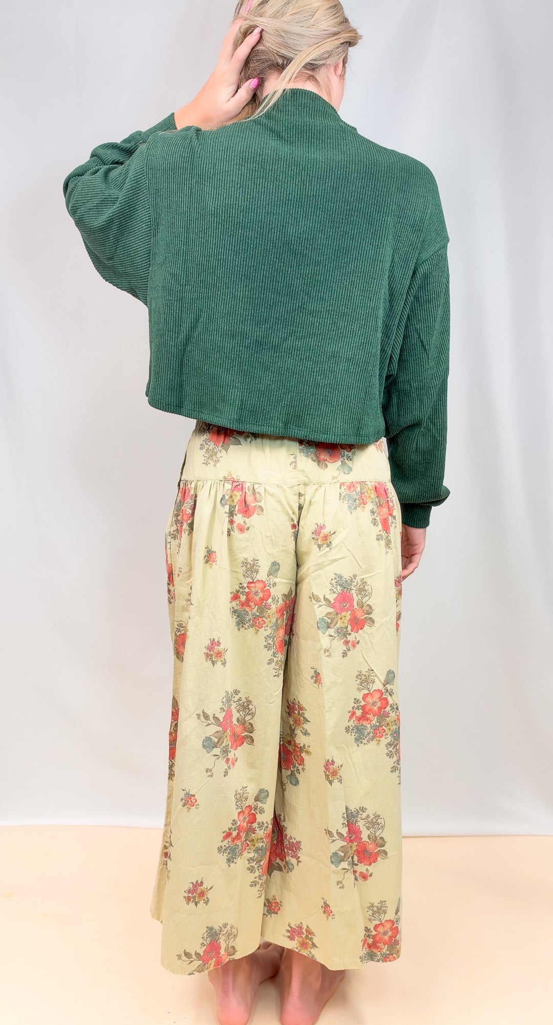 Woman shown from back view wearing green sweater and floral pants on plain background