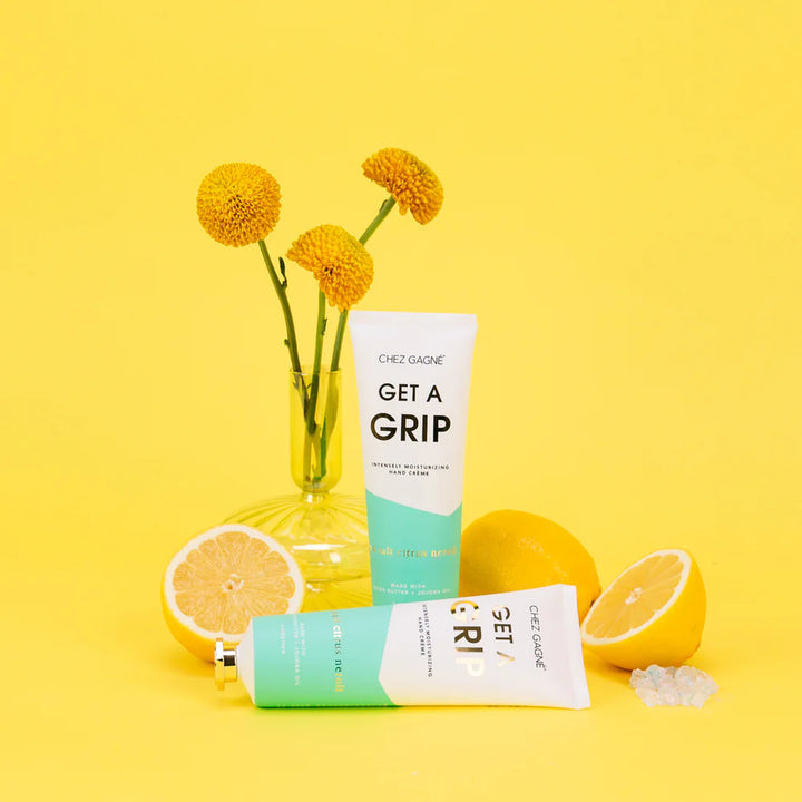 A tube and bottle of Chez Gaze Get A Grip hand cream are styled on a bright yellow background with decorative props including a glass vase with yellow flowers, whole and sliced lemons, and a few clear crystals.