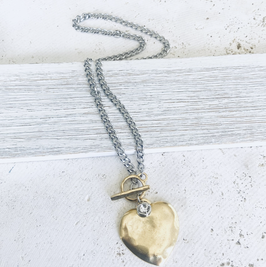 A silver chain necklace with a hammered gold heart pendant, a toggle clasp, and a small clear rhinestone charm, displayed on a white wooden surface.