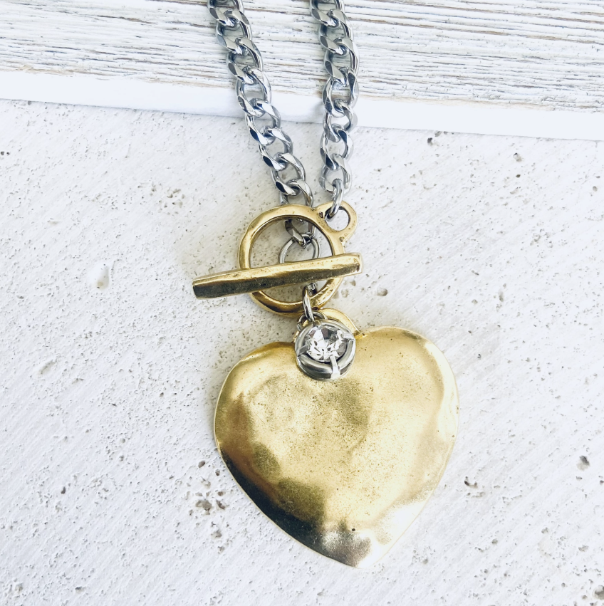 A close-up of a necklace with a hammered gold heart pendant, a small clear rhinestone charm, and a toggle clasp, hanging from a silver-toned chain on a textured white surface.