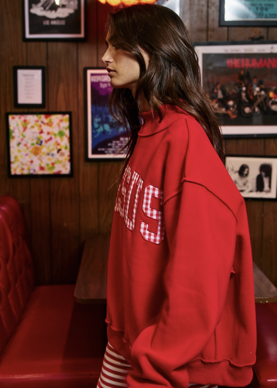 A woman in a red oversized sweatshirt with white gingham lettering stands in profile in a retro-themed room with wood-paneled walls and framed posters.