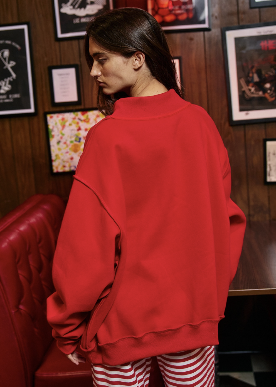 A woman wearing an oversized bright red sweatshirt and red-and-white striped pants, photographed from the back in a dimly lit indoor setting with wood-paneled walls and framed art.