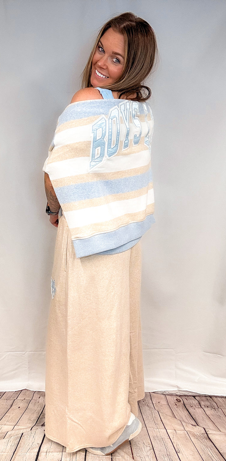 Woman shown from back wearing blue, beige and white stripe top on shoulders with beige pants on white background