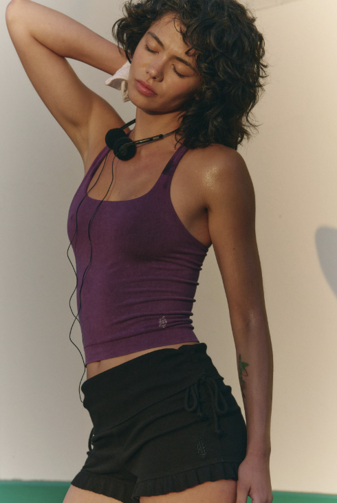 Woman wearing a purple tank top and black shorts with earphones, standing against a plain background.