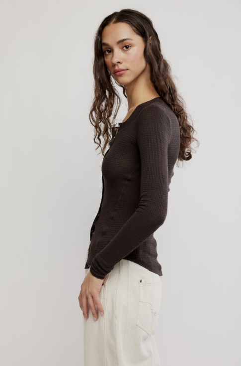 A woman wearing a dark brown ribbed long-sleeve top and off-white trousers, shown from a side profile against a light gray studio background.