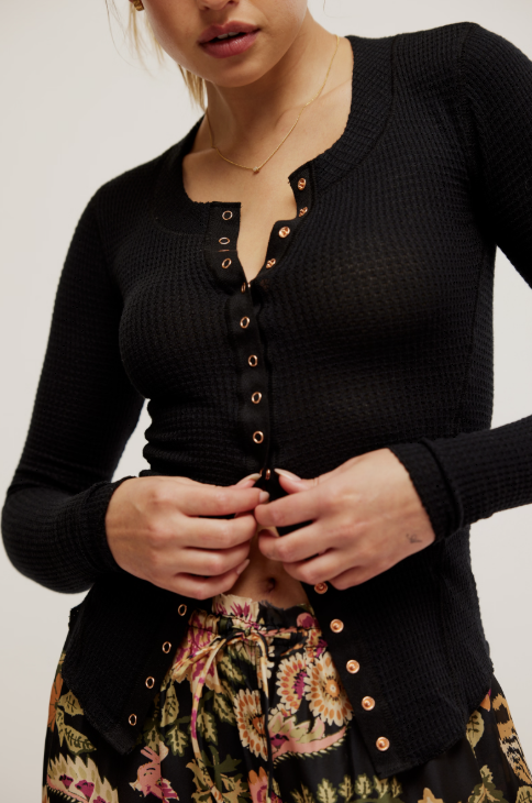 A close-up of a woman wearing a black textured long-sleeve henley cardigan with gold-tone buttons, paired with a floral skirt, against a plain light background.