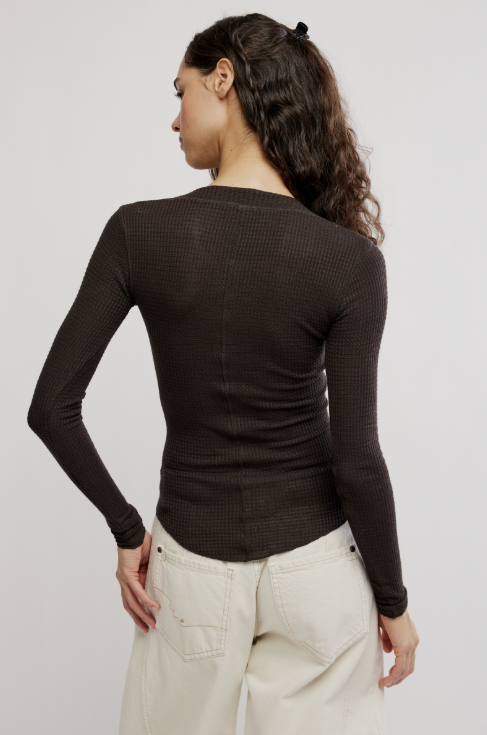 A woman shown from the back wearing a dark brown ribbed long-sleeve top and cream-colored pants, photographed against a light gray studio background.