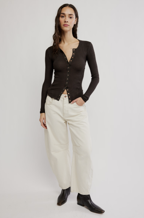 A woman modeling a dark brown long-sleeve button-front knit top with a raw hem, paired with off-white wide-leg trousers and black ankle boots, against a light gray studio background.
