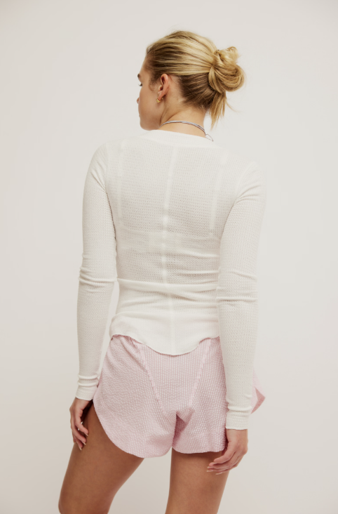 A woman shown from the back wearing a fitted white long-sleeve knit top and light pink shorts against a plain light background.
