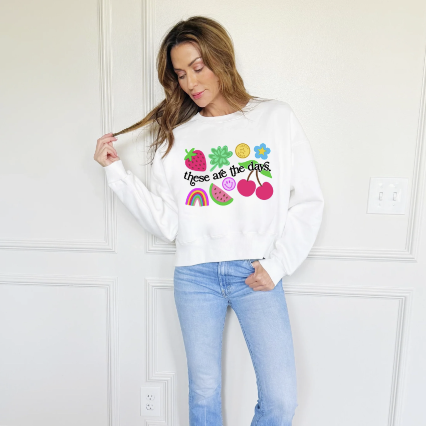 Woman wearing a white sweatshirt with colorful graphics and text, standing against a white wall.