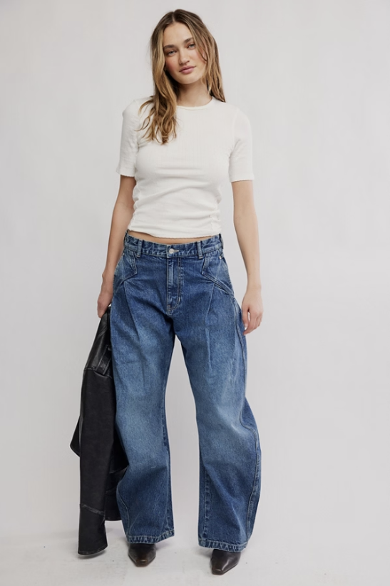 A woman modeling a pair of wide-leg blue jeans with a white short-sleeve crop top, holding a black jacket against a plain light gray studio background.
