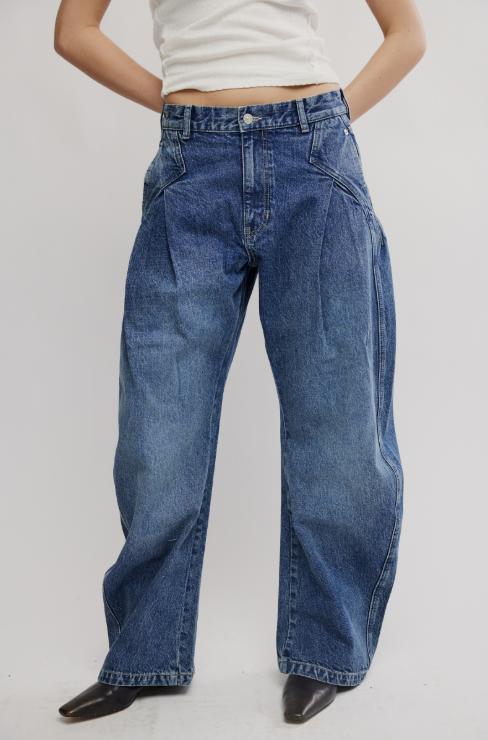 A person wearing high-waisted, wide-leg blue denim jeans with front pleats, paired with a white cropped top and black pointed shoes, shown against a light gray studio background.