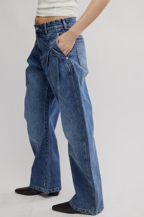 A person wearing wide-leg blue denim jeans with a high waist, pleated front, and side pockets, paired with a white cropped top and black shoes, shown against a light gray studio background.