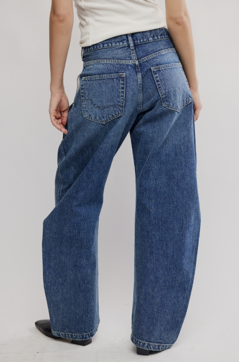 Rear view of a person wearing wide-leg blue denim jeans with a cream top and black shoes, standing against a light gray studio background.