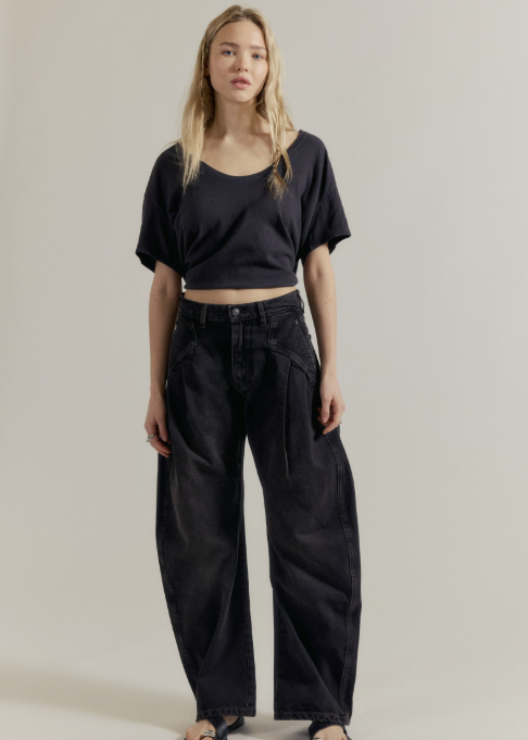 A woman stands against a plain light beige studio background wearing a loose black cropped T-shirt, very dark wide-leg jeans, and black sandals.