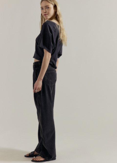 A woman stands in profile wearing a loose short-sleeve dark top, dark wide-leg jeans, and black sandals against a plain light gray studio background.
