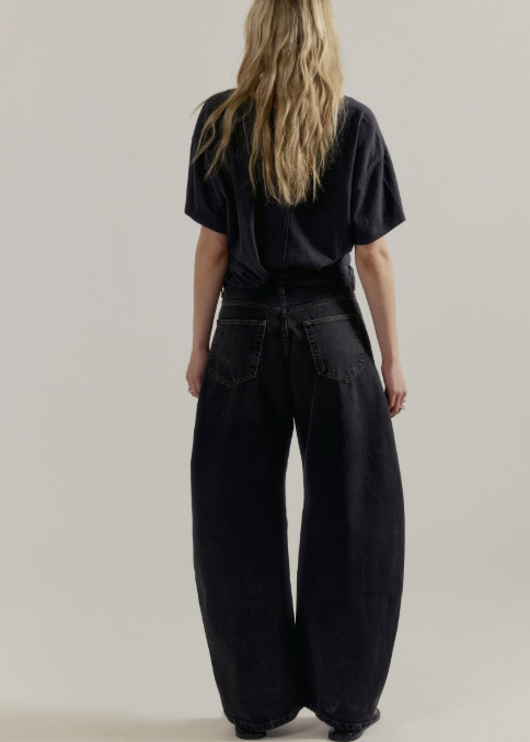 A woman viewed from behind wearing a black short-sleeve t-shirt and very wide-leg black denim pants, standing against a plain light gray studio background.
