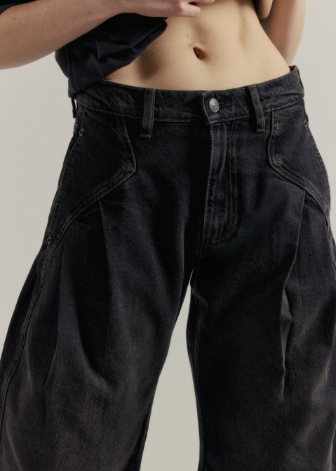Close-up of a person wearing oversized black denim jeans with a cropped black top, shown against a plain light gray studio background.