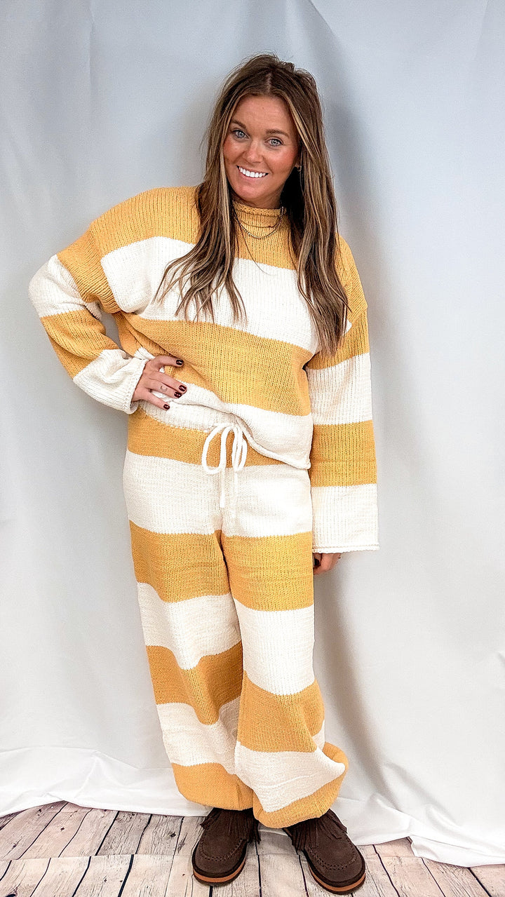 A woman modeling a matching knit lounge set with wide horizontal stripes in mustard yellow and cream, featuring a long-sleeve top and drawstring pants, paired with brown slip-on shoes against a white draped studio backdrop.