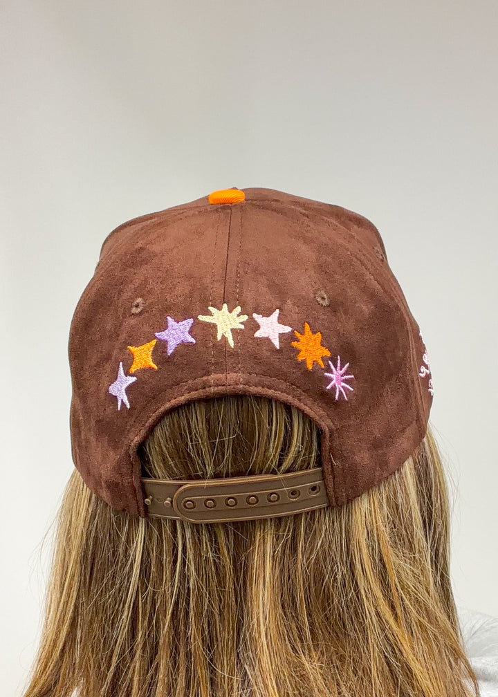 Woman wearing brown faux suede hat with stars embroidered across back on plain background