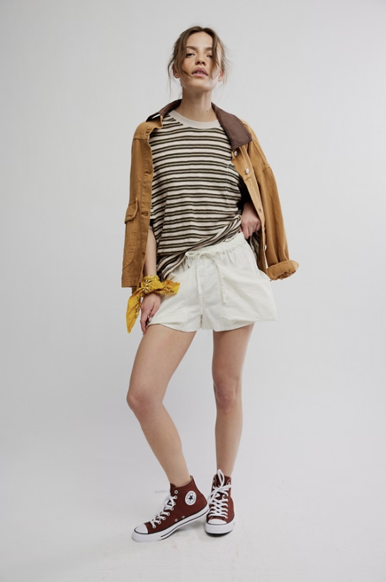 Woman wearing a striped shirt, beige jacket, white shorts, and brown sneakers on a plain background