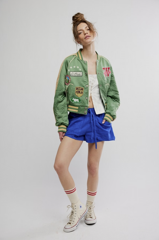 Person wearing a green bomber jacket with patches, blue shorts, and white sneakers on a plain background