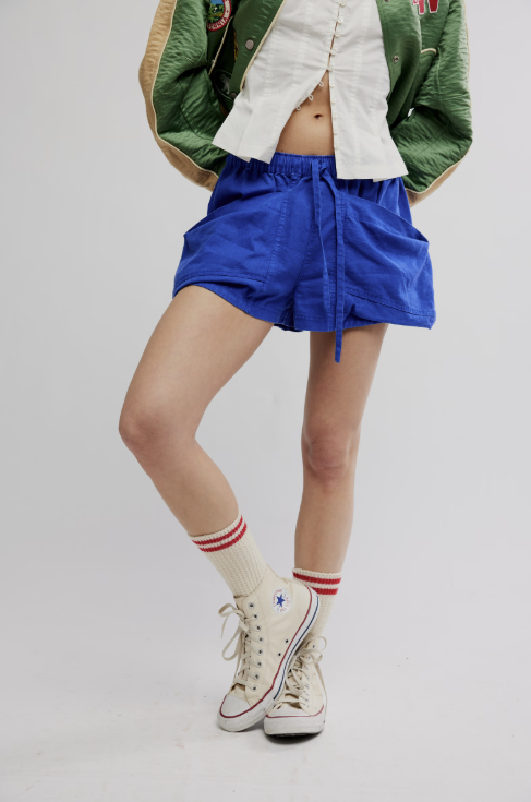 Person wearing blue shorts, a green jacket, and white sneakers with red stripes on a plain background