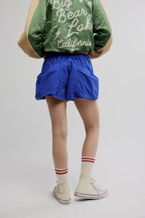 Person wearing a green jacket with text, blue shorts, and white sneakers on a plain background