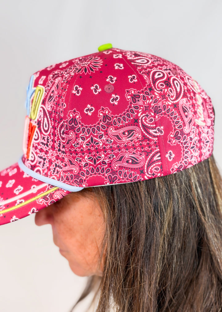 Woman shown from side view wearing maroon bandana print trucker hat with lime top button on plain background