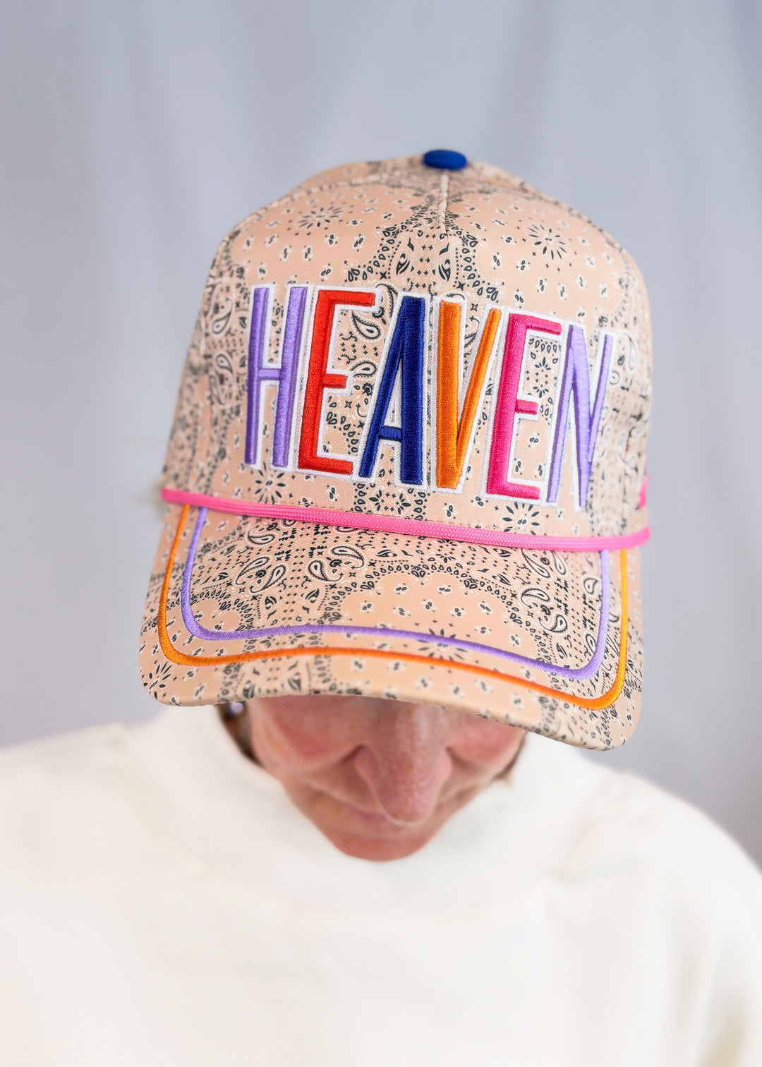Woman wearing beige bandana print trucker hat with 'HEAVEN' embroidered across front on plain background