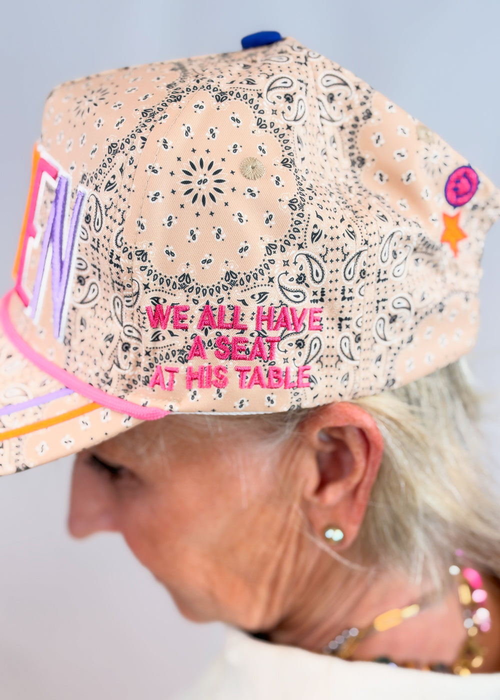Woman shown from side wearing tan bandana print trucker hate with 'WE ALL HAVE A SEAT AT HIS TABLE' embroidered on side on plain background