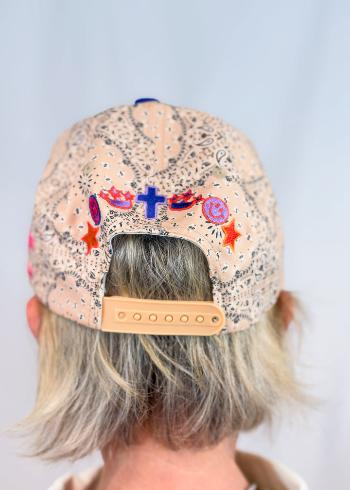 Woman shown from back wearing tan bandana print trucker hat with multiple spiritual embroidery designs on back on plain background