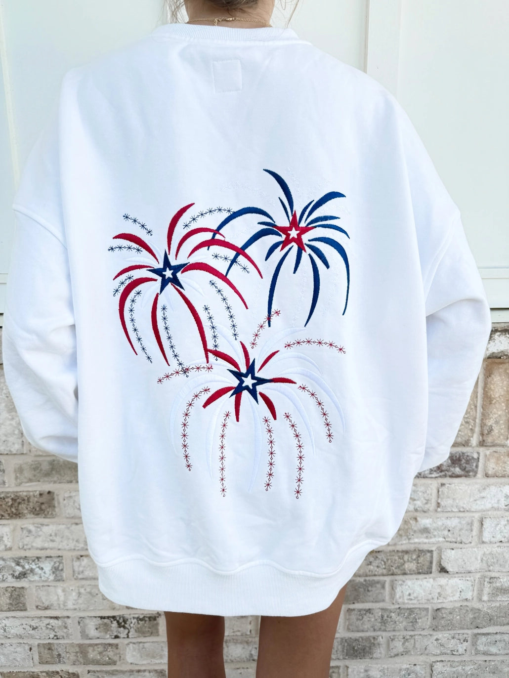 White sweatshirt with red, white, and blue fireworks design on a person standing against a brick wall.