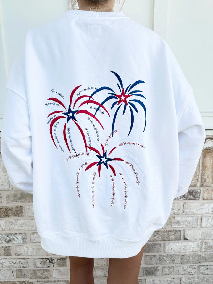 White sweatshirt with red, white, and blue fireworks design on a person standing against a brick wall.