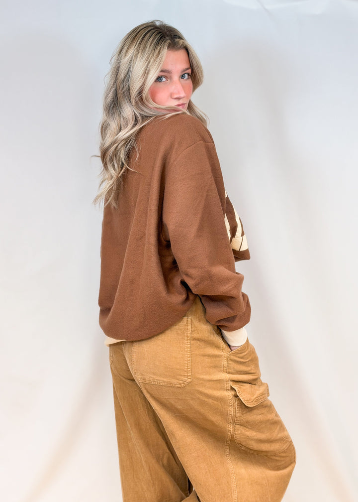 Woman shown from side view wearing brown mock neck sweatshirt and brown pants on plain background