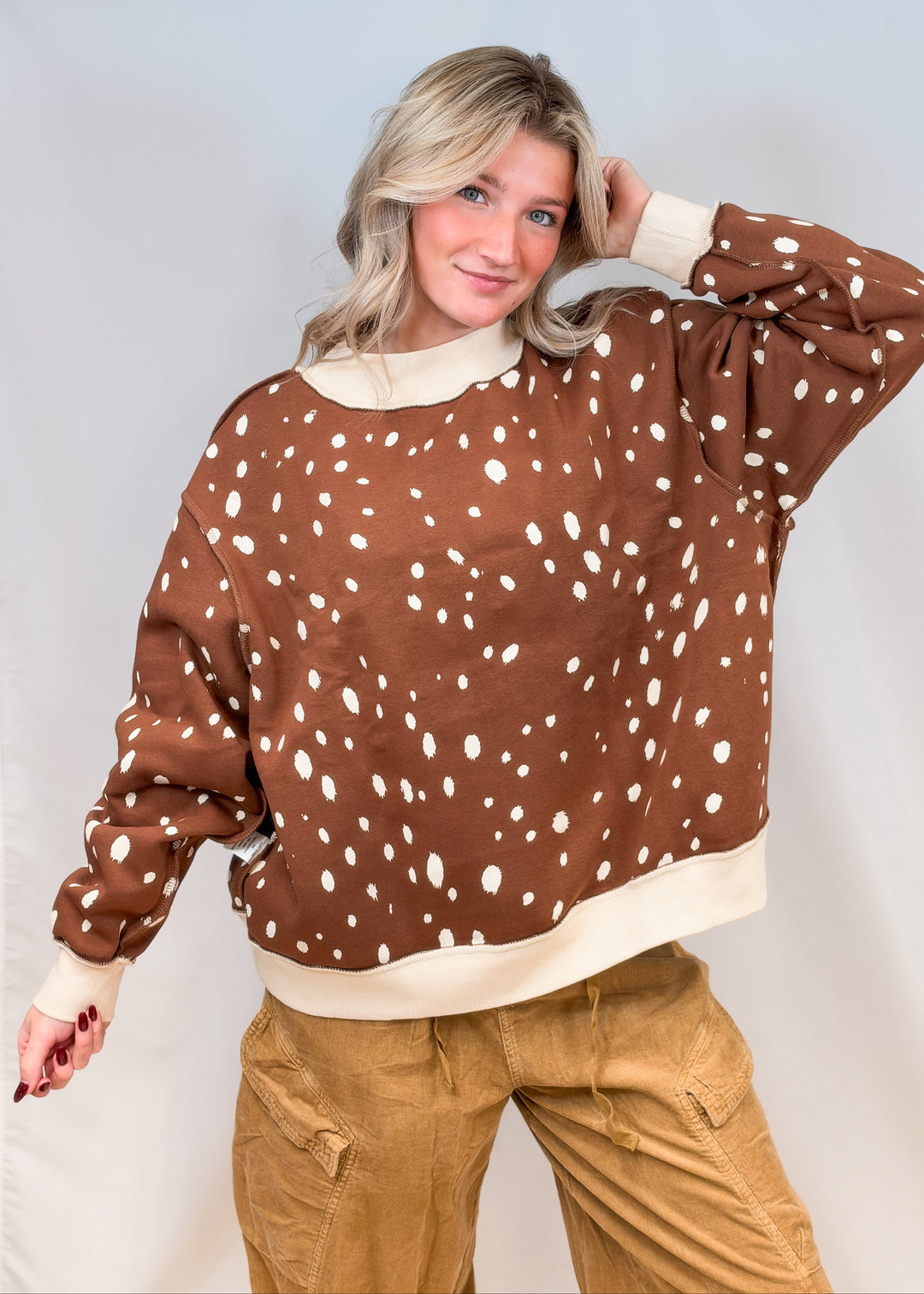 Woman wearing brown mock neck sweatshirt with cream polka dots and brown pants on plain background