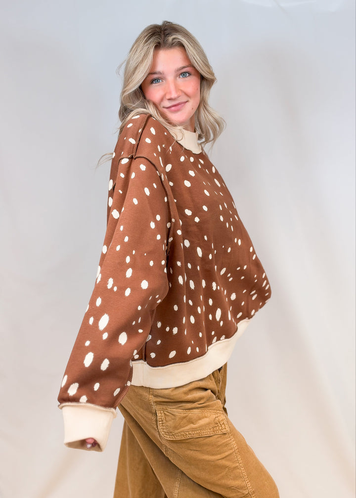 Woman shown from side view wearing brown mock neck sweatshirt with cream polka dots and brown pants on plain background