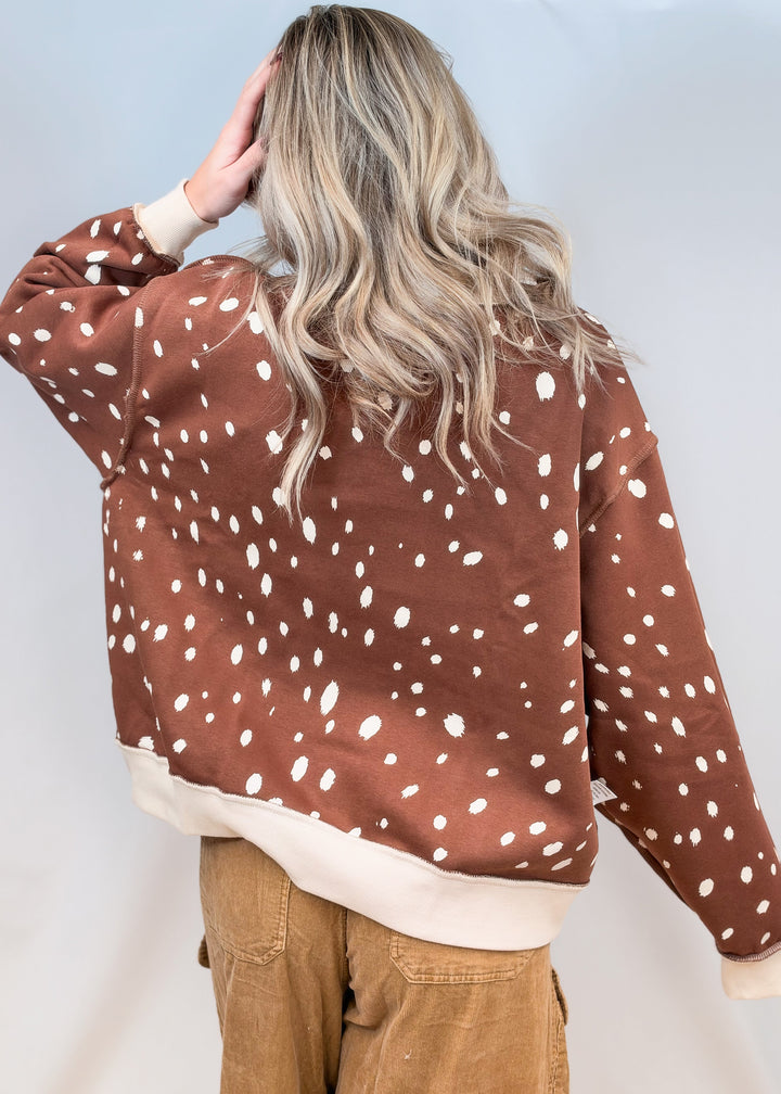Woman shown from back view wearing brown mock neck sweatshirt with cream polka dots and brown pants on plain background