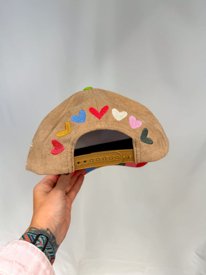 Brown suede trucker hat shown from back with colorful hearts embroidered across back on white background