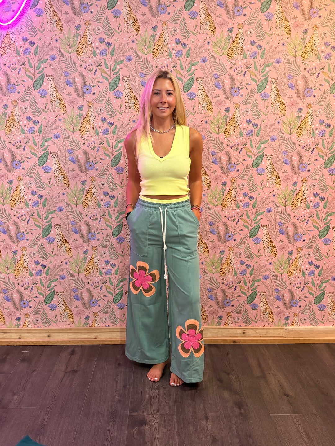 Woman in a yellow top and green pants with floral designs standing against a patterned wall.
