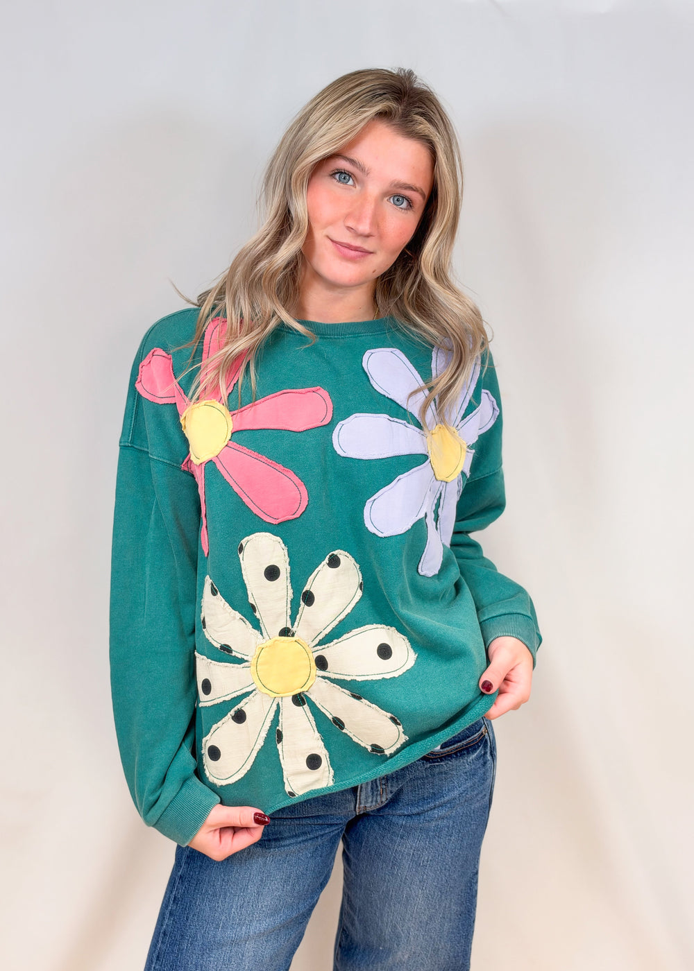 Woman wearing teal crewneck with applique flowers and jeans on plain background