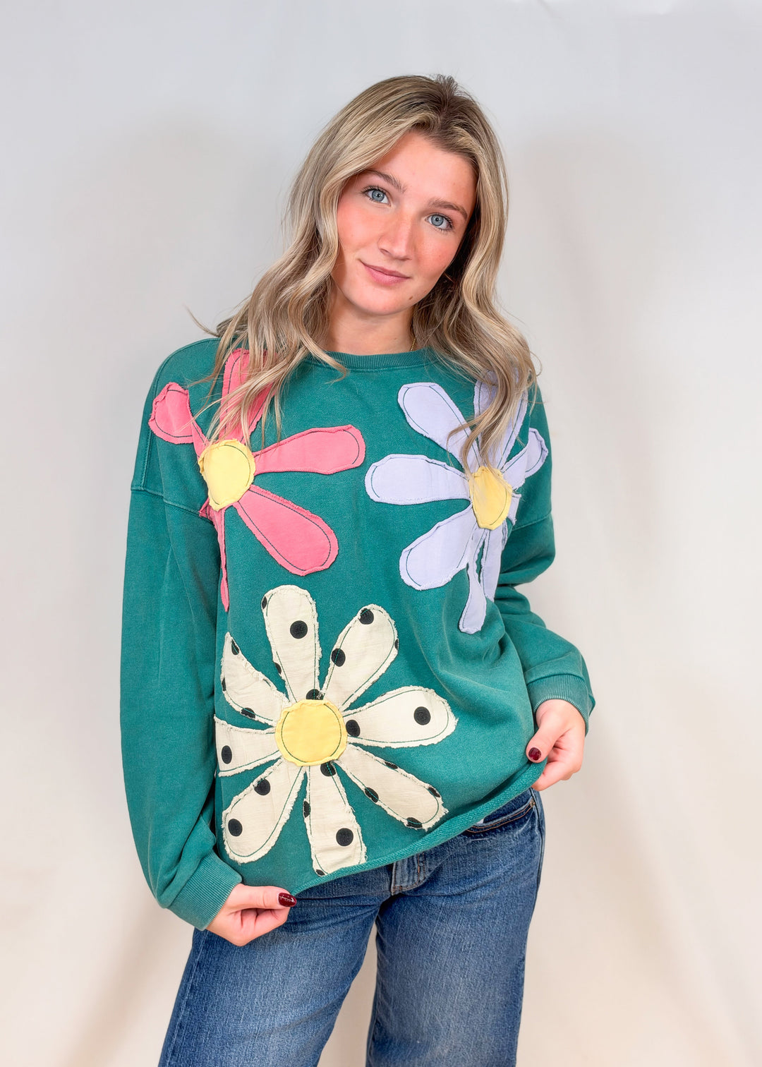 Woman wearing teal crewneck with applique flowers and jeans on plain background