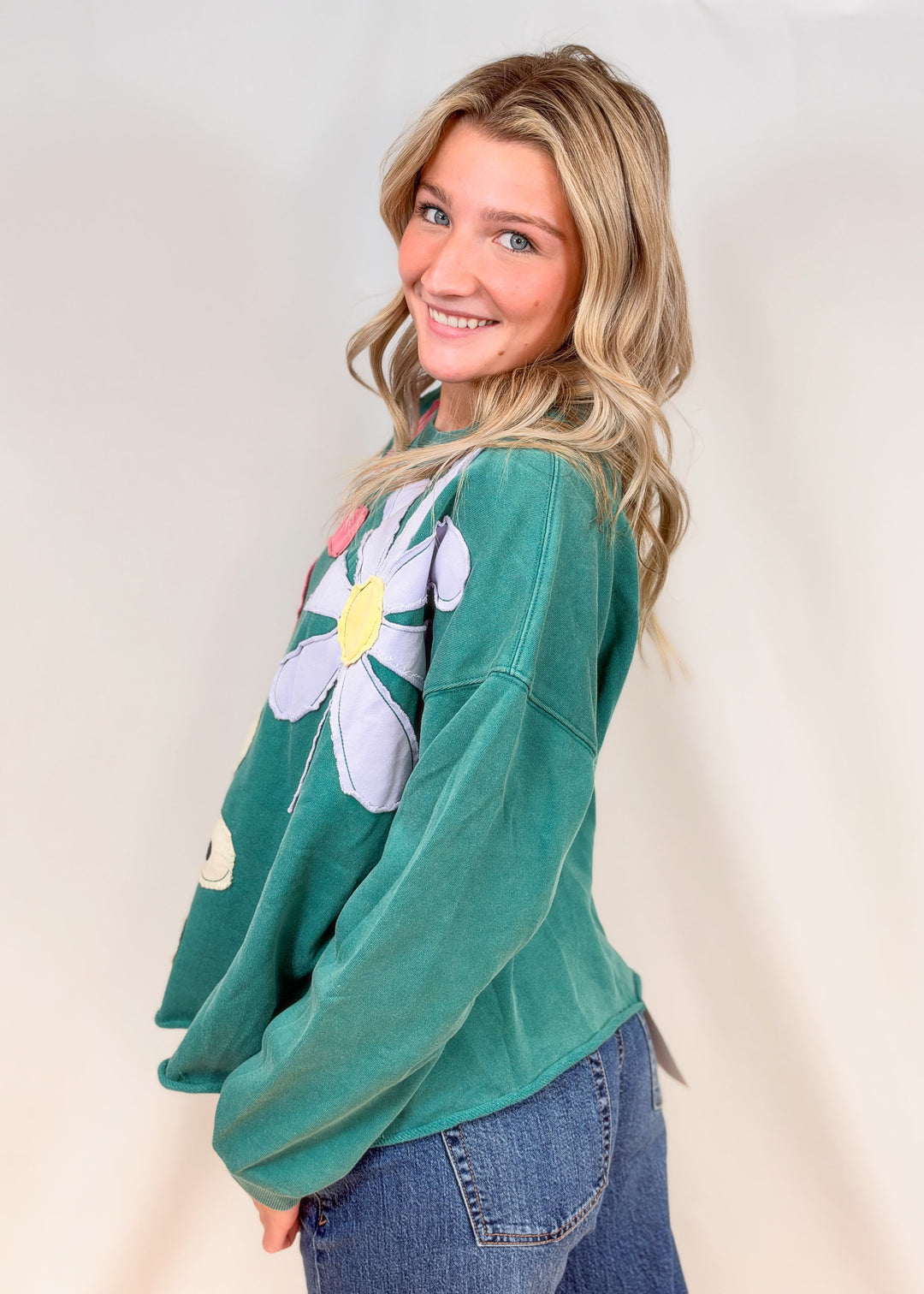 Woman shown from side view wearing teal crewneck with applique flowers and jeans on plain background