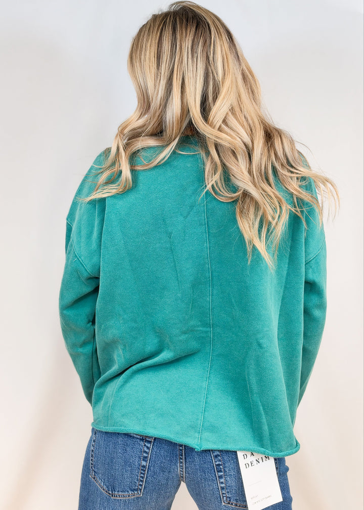 Woman shown from back view wearing teal crewneck and jeans on plain background